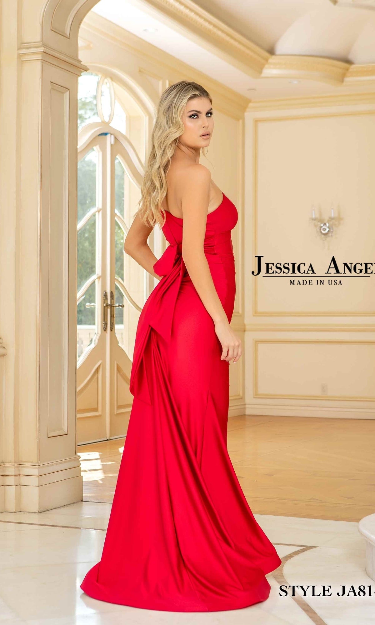 A woman in a Jessica Angel 814 One-Shoulder Prom Gown—a striking floor-length red dress—stands in an elegant room with cream walls and large windows.