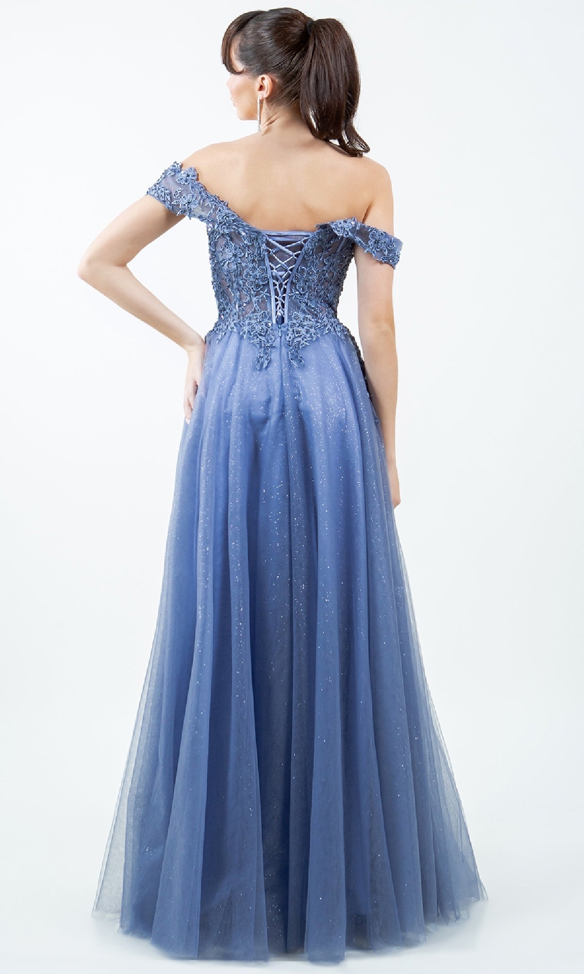 Woman in the Cinderella Couture 8152J long prom ball gown with an off-the-shoulder lace-up back and glittery tulle skirt, standing against a plain light background, facing away from the camera.