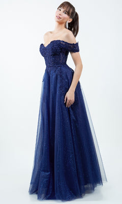 A woman wears the Cinderella Couture 8152J Long Prom Ball Gown, a navy blue off-the-shoulder dress with a floor-length tulle skirt and sequin embellishments, standing against a plain light background.