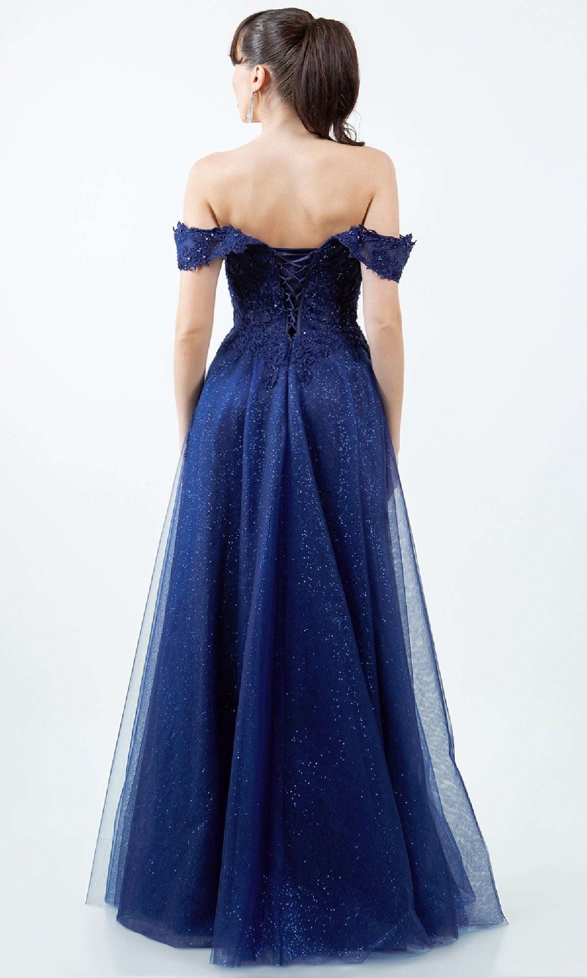A woman wears the Cinderella Couture 8152J long prom ball gown in navy blue, featuring an off-the-shoulder neckline, lace-up corset back, and a floor-length glittery tulle skirt as she stands facing away.