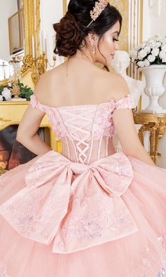 A woman wearing the Cinderella Couture 8160J quinceanera dress, an ornate pink off-the-shoulder ball gown with embroidered lace appliques and a large bow, stands showing its lace-up back in a room decorated with gold and flowers.