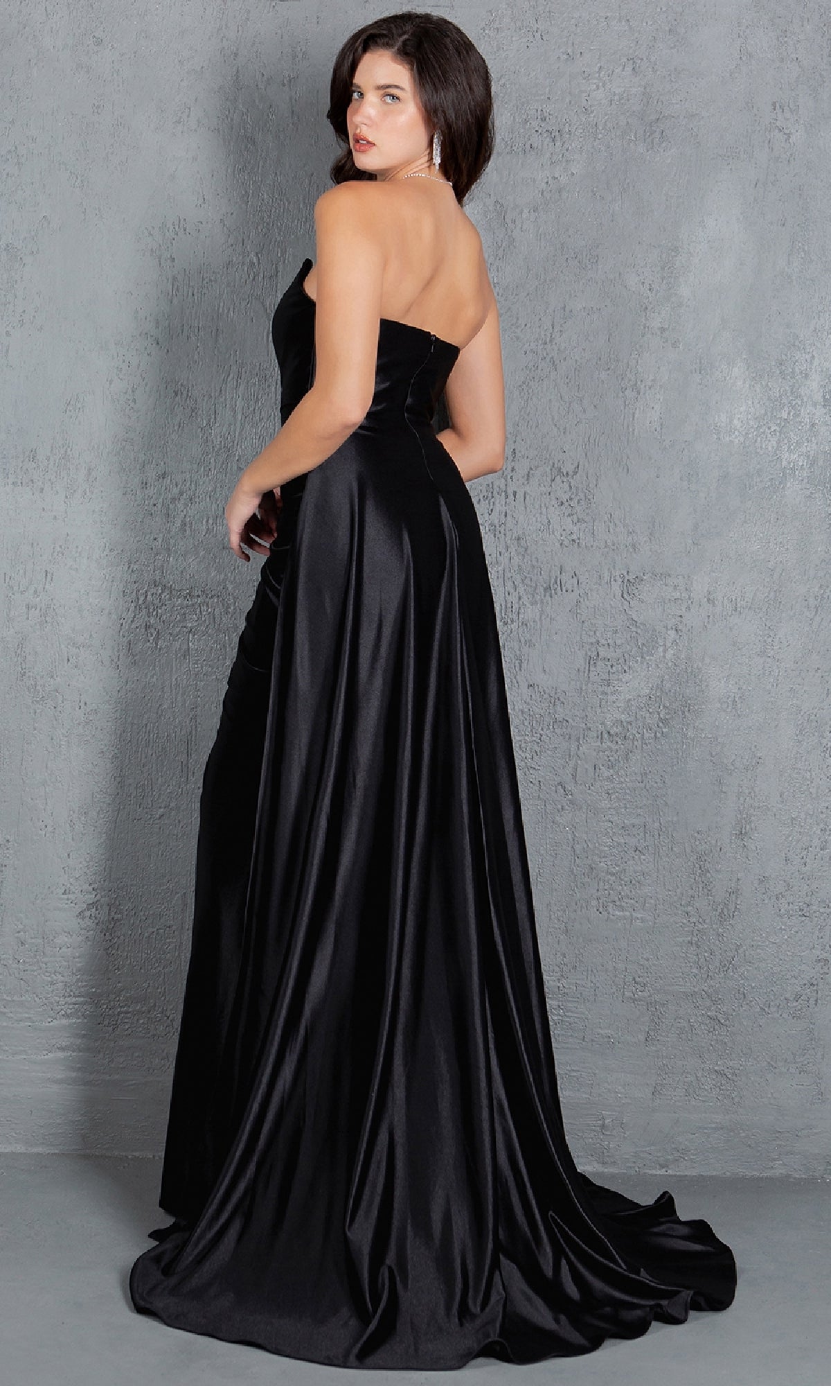 A woman wearing the Cinderella Couture 8161J Strapless Long Prom Dress stands with her back turned, glancing over her shoulder against a textured gray background.