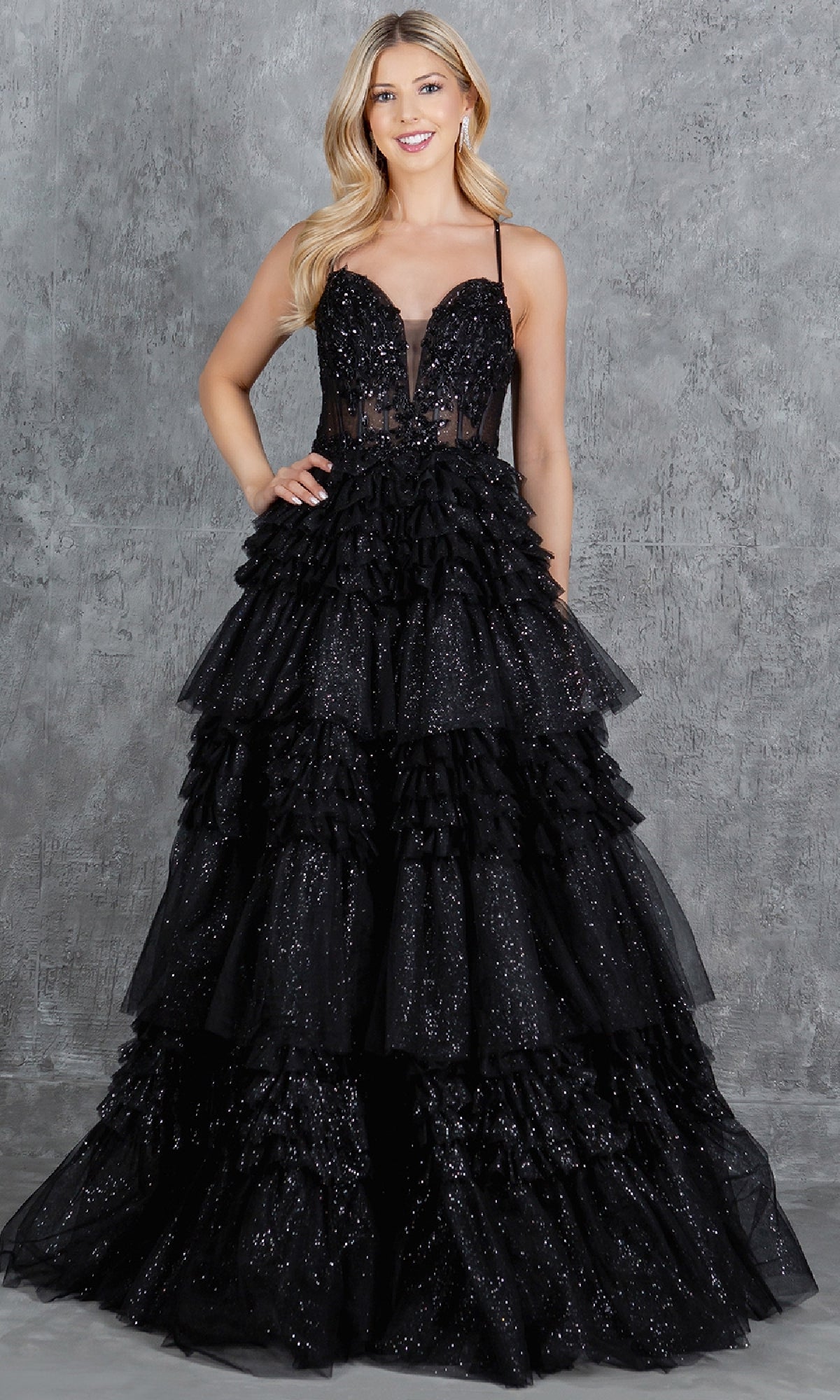 A woman poses in front of a gray textured background wearing the Glitter Prom Ball Gown: Cinderella Couture 8163J, a sleeveless black dress with a beaded corset bodice and tiered ruffled floor-length skirt.