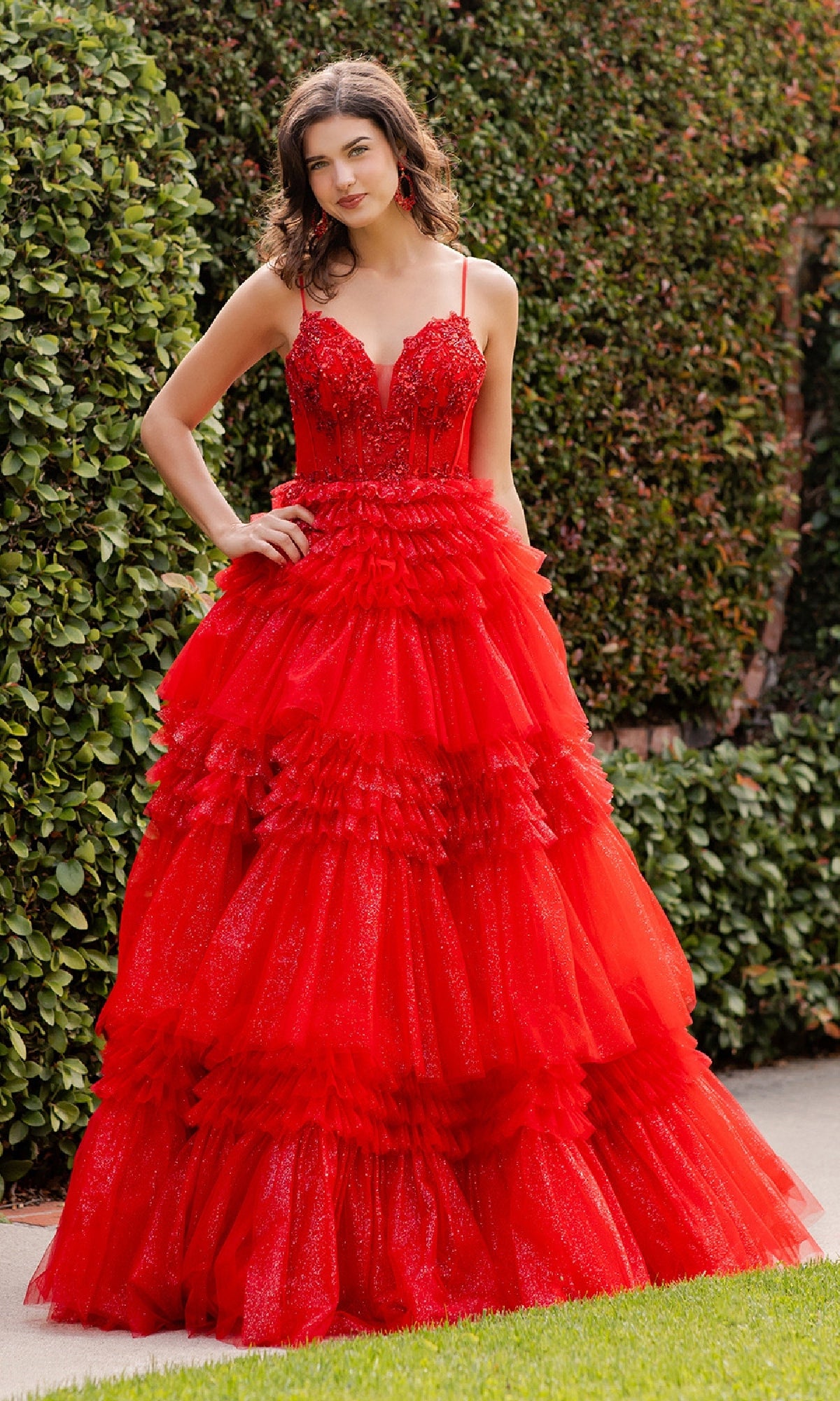 A woman stands outdoors in front of green hedges, wearing the Glitter Prom Ball Gown: Cinderella Couture 8163J—a vibrant red, floor-length tulle gown with a beaded corset bodice and fitted, embellished details.