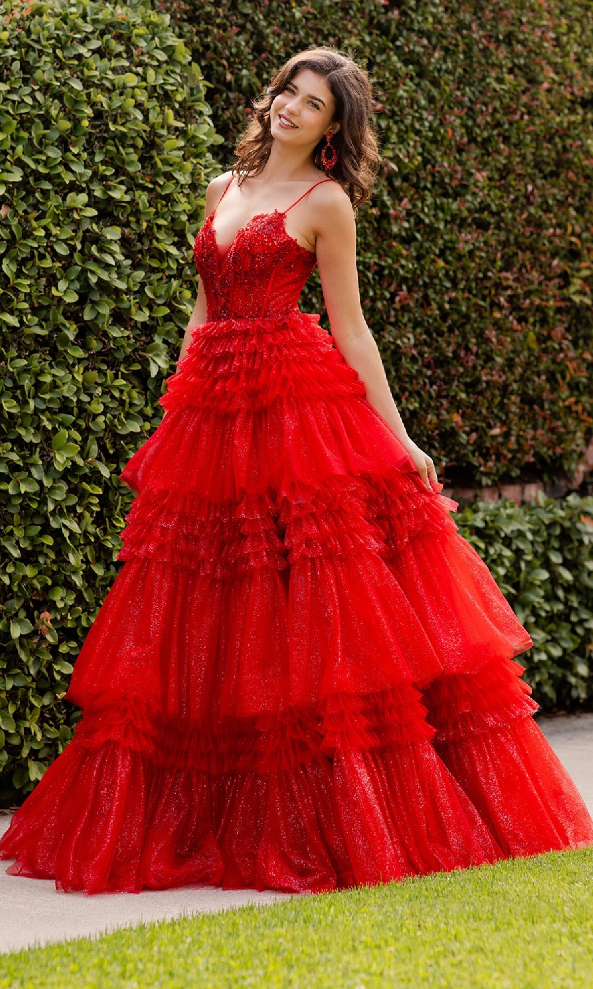 A woman stands outdoors before greenery, wearing the Glitter Prom Ball Gown: Cinderella Couture 8163J—a bright red dress with a tiered skirt, spaghetti straps, and a beaded corset bodice in a glittering A-line style.