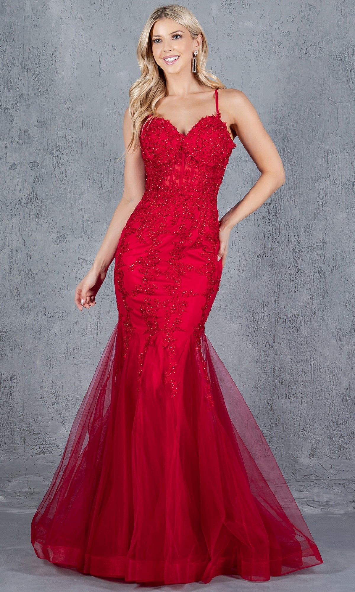 A woman wearing the Lace-Up Long Prom Dress: Cinderella Couture 8175J, a fitted red gown with beaded details and a tulle mermaid skirt, stands against a textured gray background.
