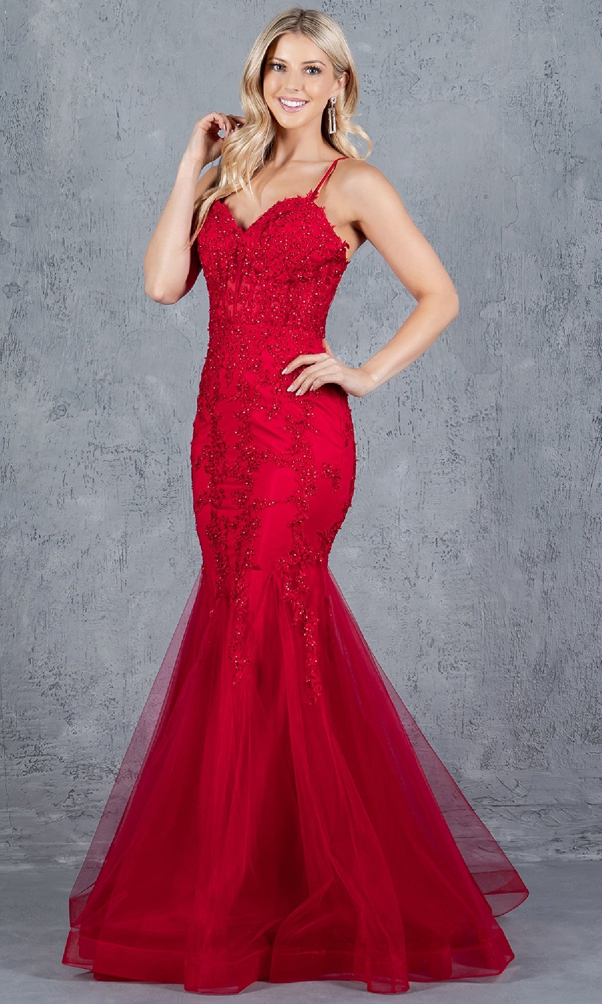 A woman poses confidently before a gray textured backdrop in the Lace-Up Long Prom Dress: Cinderella Couture 8175J, a red evening gown with beaded lace applique and thin straps, one hand on her hip and the other touching her hair.
