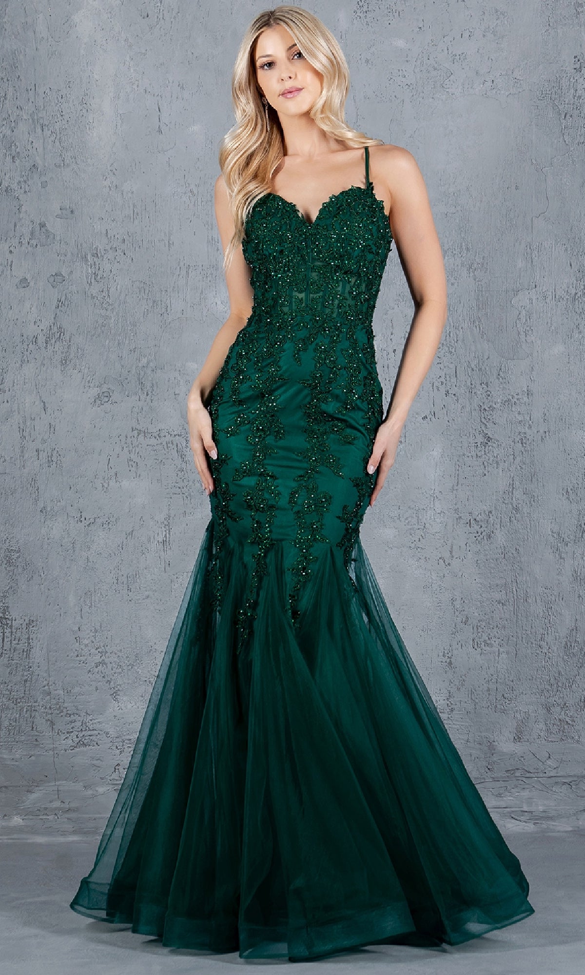 A woman models the Cinderella Couture 8175J, a sleeveless dark green lace-up mermaid prom dress with beaded detailing, standing against a textured gray background.