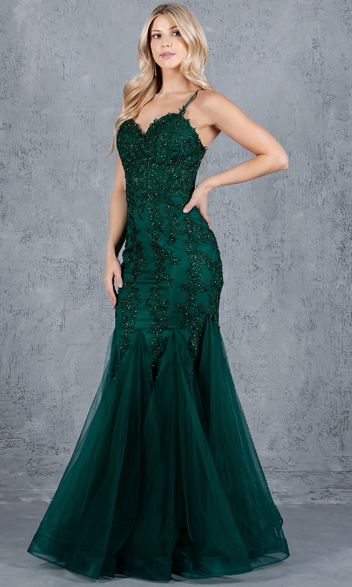 A woman stands before a gray textured wall in the Lace-Up Long Prom Dress: Cinderella Couture 8175J—a dark green mermaid gown with beaded accents and tulle at the hem.