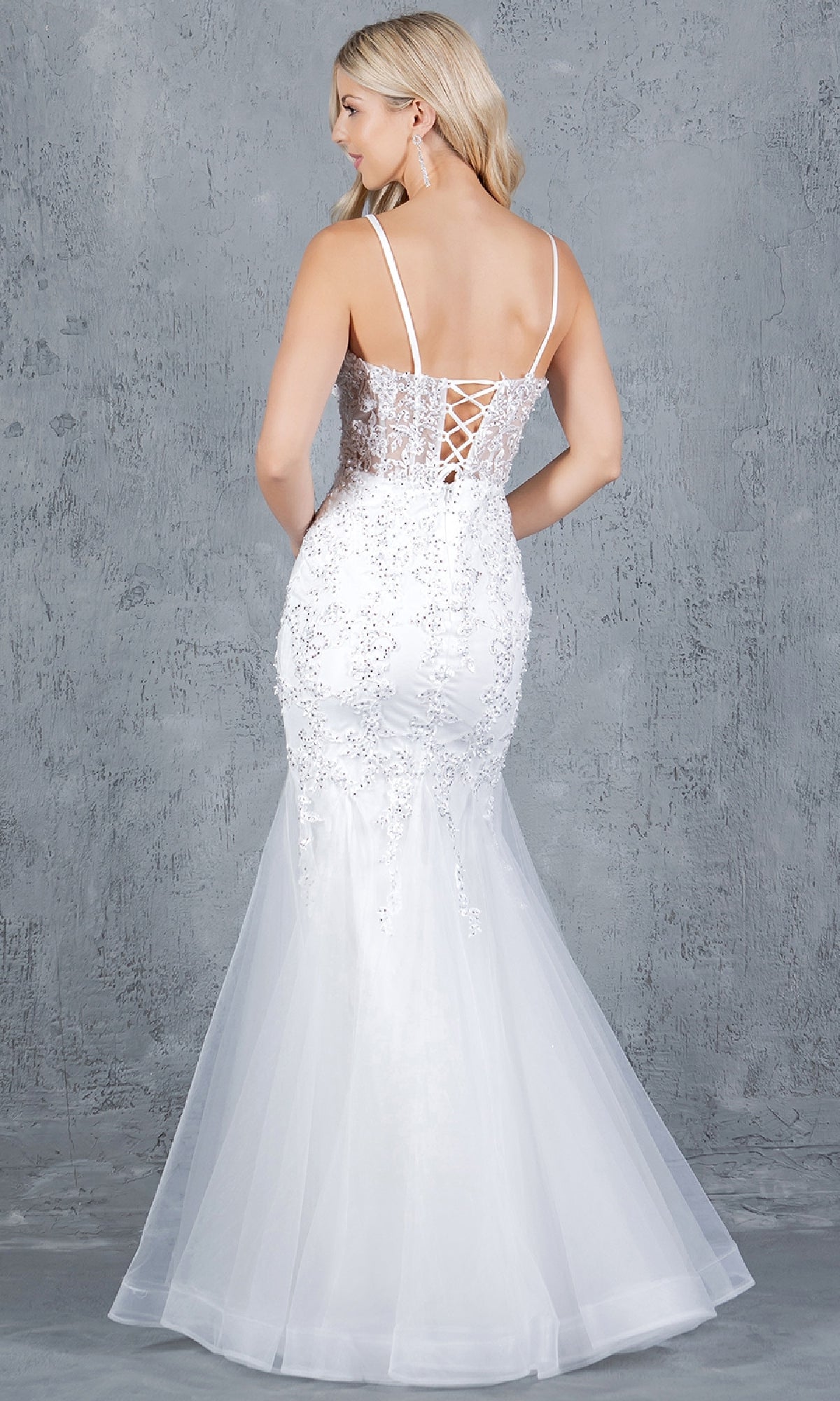 A woman stands facing away against a textured gray background wearing the Cinderella Couture 8175J—a sleeveless, white, fitted lace-up long prom dress with beaded lace appliqué and a tulle mermaid skirt.
