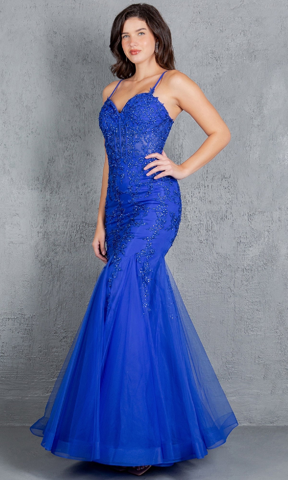 A woman stands against a gray textured background wearing the Lace-Up Long Prom Dress: Cinderella Couture 8175J, a fitted bright blue mermaid gown with beaded lace appliqué and thin straps.