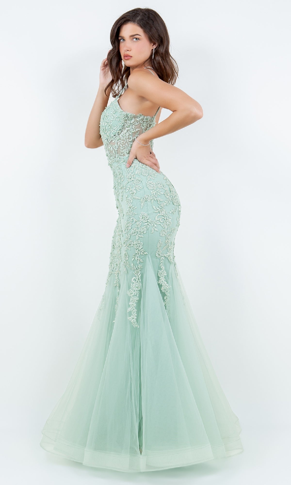 A woman poses sideways against a plain white background in the Lace-Up Long Prom Dress: Cinderella Couture 8175J, featuring a fitted light green mermaid silhouette with beaded lace applique and a flared tulle skirt.