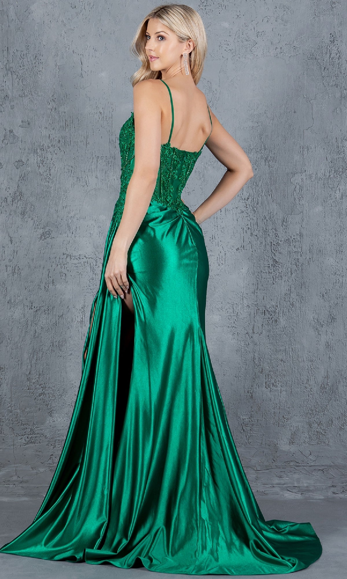 A woman poses against a gray textured backdrop in the Long Corset Prom Dress: Cinderella Couture 8177J, featuring a shiny emerald green finish, thin straps, and a high slit, as she looks over her shoulder.