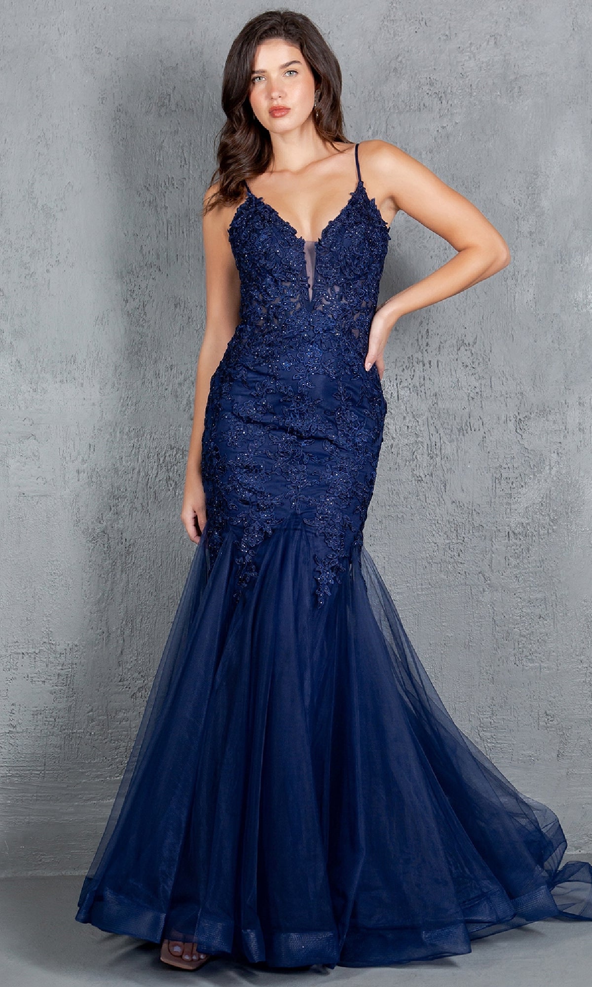 A woman stands against a textured gray backdrop wearing the Beaded Long Prom Dress: Cinderella Couture 8178J, a dark blue sleeveless mermaid gown with beaded embroidery, lace, and tulle details.