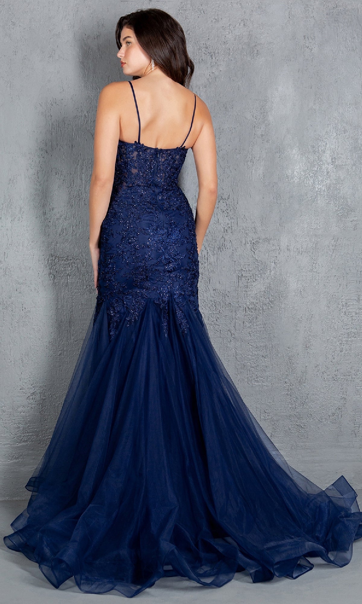 A woman with long dark hair models the Cinderella Couture 8178J, a navy blue beaded long prom dress with spaghetti straps and mermaid silhouette, standing against a gray textured wall and facing away from the camera.