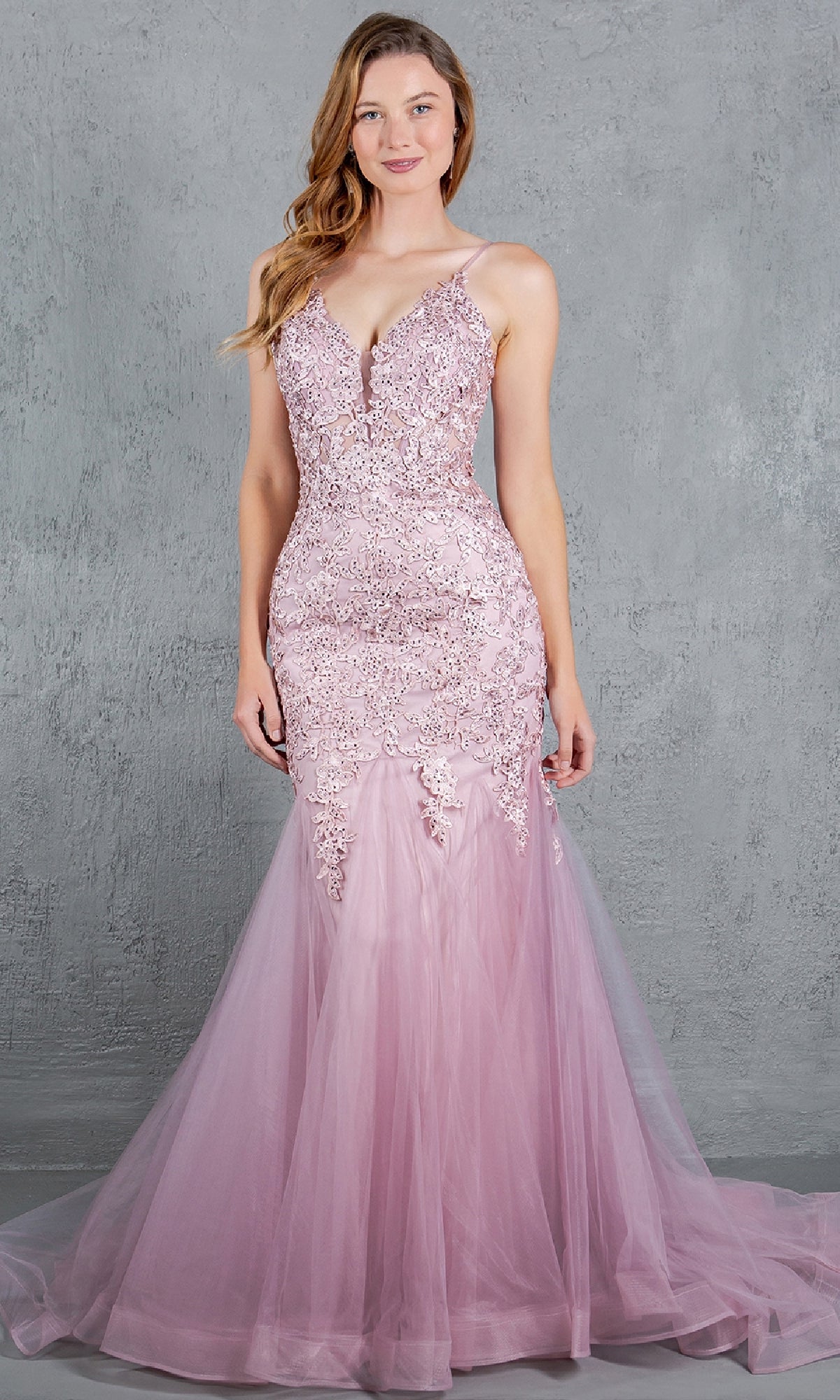 A woman in the Cinderella Couture 8178J Beaded Long Prom Dress—sleeveless, floor-length, pink with lace and tulle and delicate beaded embroidery—stands before a gray textured backdrop.
