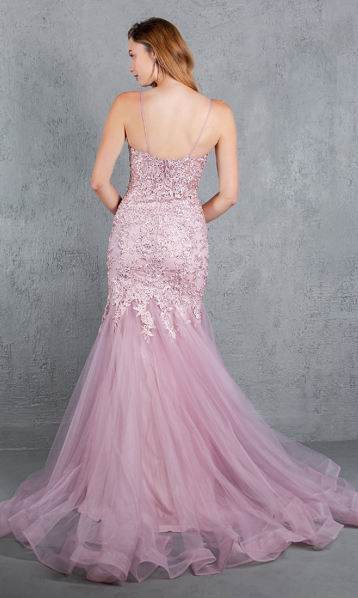 A woman wearing the Beaded Long Prom Dress: Cinderella Couture 8178J in light purple, featuring lace detail, beaded embroidery, and thin straps, stands with her back to the camera against a plain gray wall.