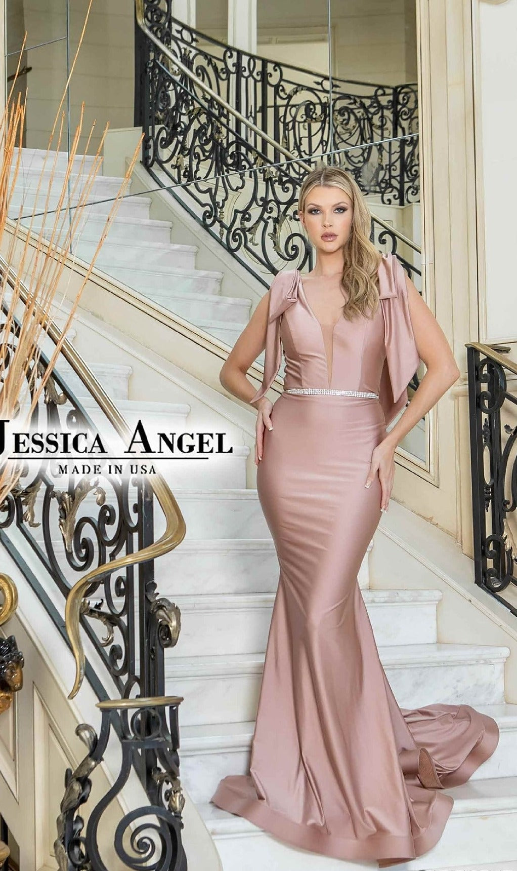 A woman wears the Jessica Angel 819 Shoulder-Bows Long Prom Dress in blush pink, featuring a fitted mermaid silhouette, deep v-neck, and statement shoulder bows as she stands on a white staircase with black and gold railings.