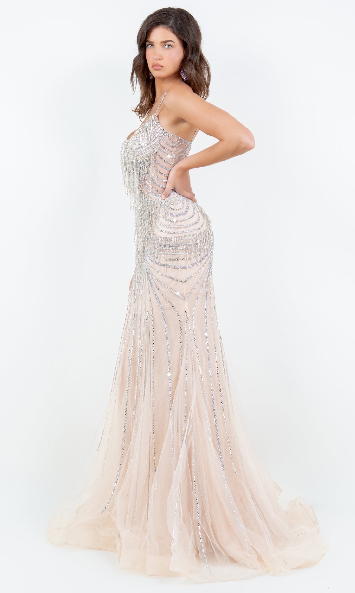 A woman stands in profile against a white background, wearing the Cinderella Couture 8192J—a floor-length, sleeveless prom dress adorned with silver beading and shimmering sequins.