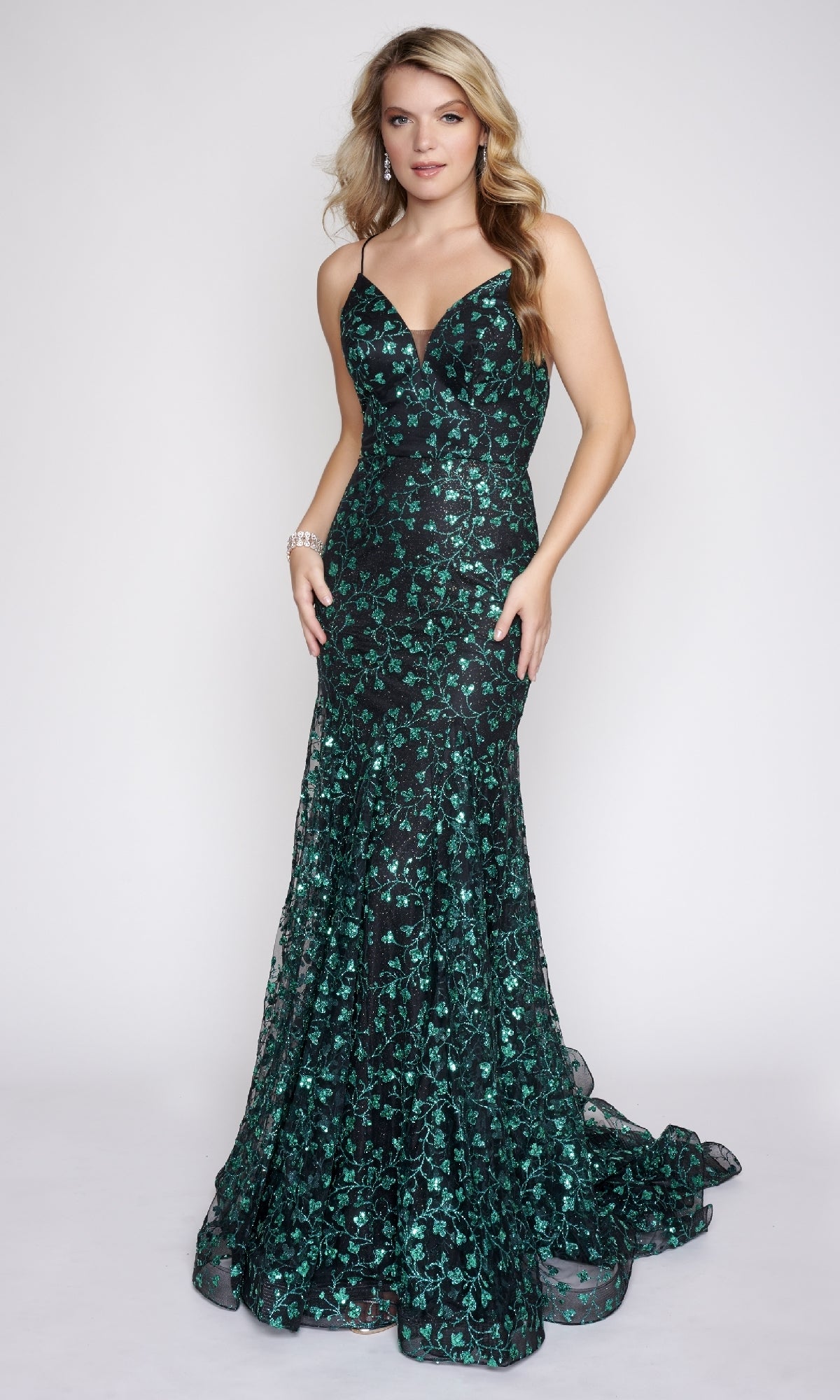 A woman poses against a plain light background in the Long Formal Dress: Nina Canacci 8210—a fitted, dark green and black evening gown with floral embellishments, thin straps, and an adjustable back tie.