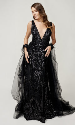 A woman wears the Nina Canacci 8224 long formal dress—a sleeveless, floor-length black gown with a deep V-neck, sequin embellishments, and tulle accents—standing against a plain light background.