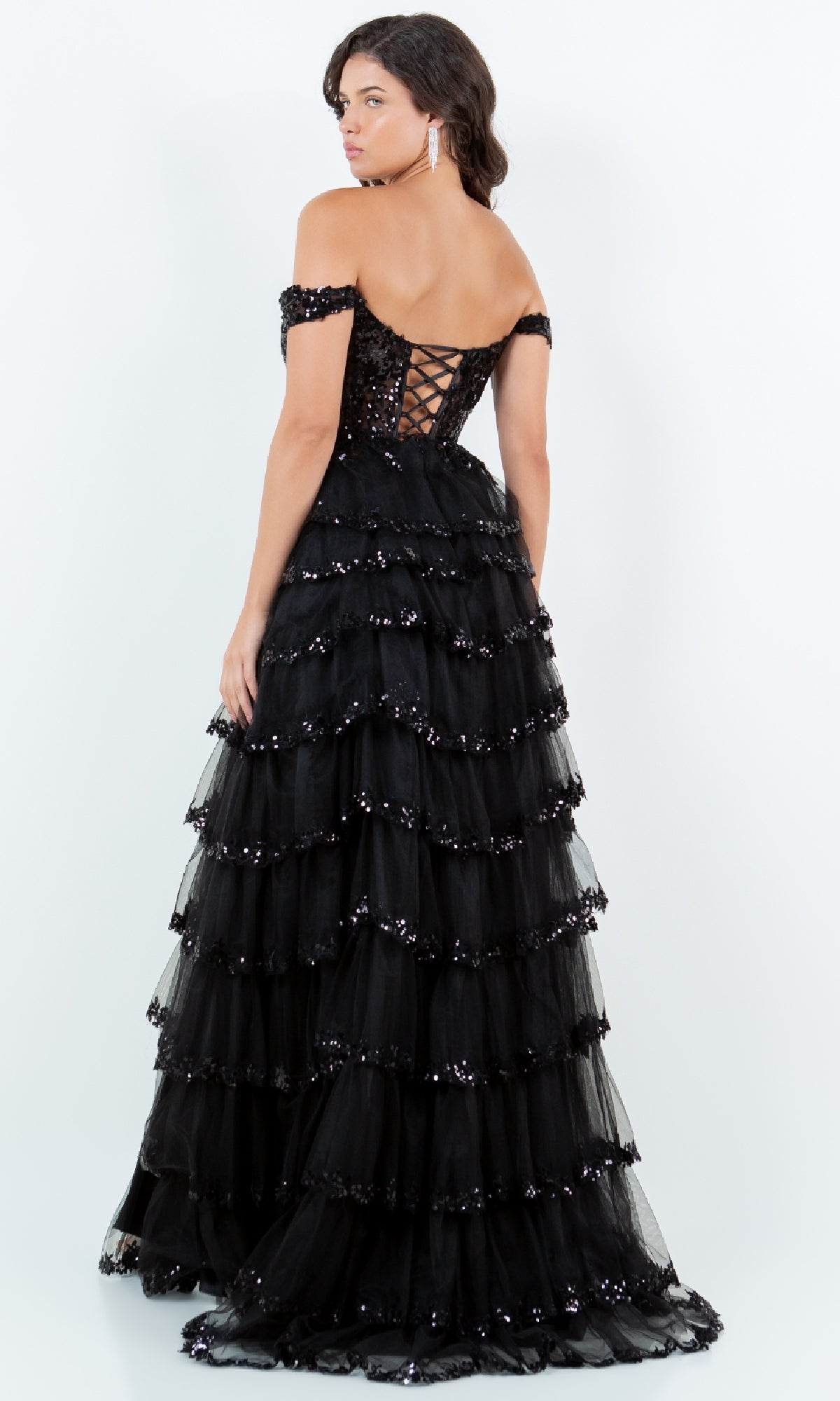 A woman in the Cinderella Couture 8228J Ruffle Prom Ball Gown with layered ruffles, sequin embroidery, and a lace-up back stands facing away from the camera.