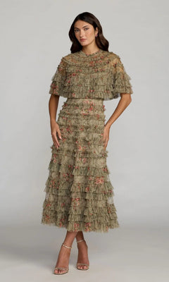 A woman stands against a plain grey background wearing the Mac Duggal 8231 tea-length party dress in olive, featuring floral details and short sleeves, paired with nude heeled sandals.