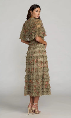 A woman stands in profile, wearing the Mac Duggal 8231 Tea-Length Party Dress—a calf-length, olive-green dress featuring tiered lace and delicate floral embroidery—paired with nude high-heeled sandals against a plain light gray background.