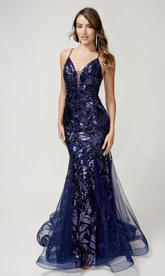 A woman stands against a plain light background wearing the Nina Canacci 8236, a sleeveless navy blue long formal mermaid dress with sequins and a sheer overlay.