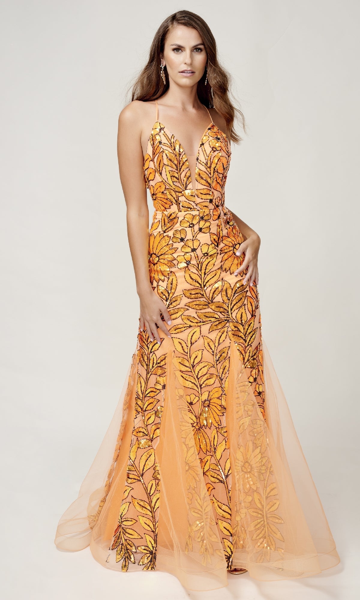 Woman in the Long Formal Dress: Nina Canacci 8236—sleeveless, floor-length, orange with a deep V-neckline and intricate floral patterns—stands against a plain light background.