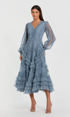 A woman stands against a plain light background wearing the Mac Duggal 8247 blue midi wedding guest dress with ruffled tiers and sheer long sleeves, styled with strappy heels.