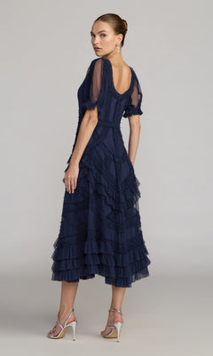 A woman stands sideways in the Mac Duggal 8248 Midi Wedding Guest Dress—a navy blue, short-sleeve, ruffled midi dress—paired with silver high-heeled sandals, glancing over her shoulder against a plain background.