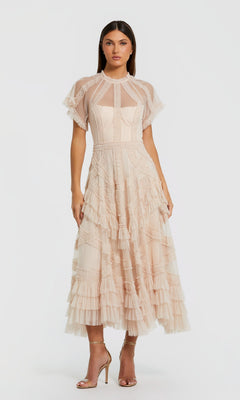 A woman wearing the Mac Duggal 8249 Tea-Length Party Dress in blush—featuring short sleeves, ruffle details, and sheer panels—stands against a plain white background in gold high-heeled sandals, embodying effortless elegance.