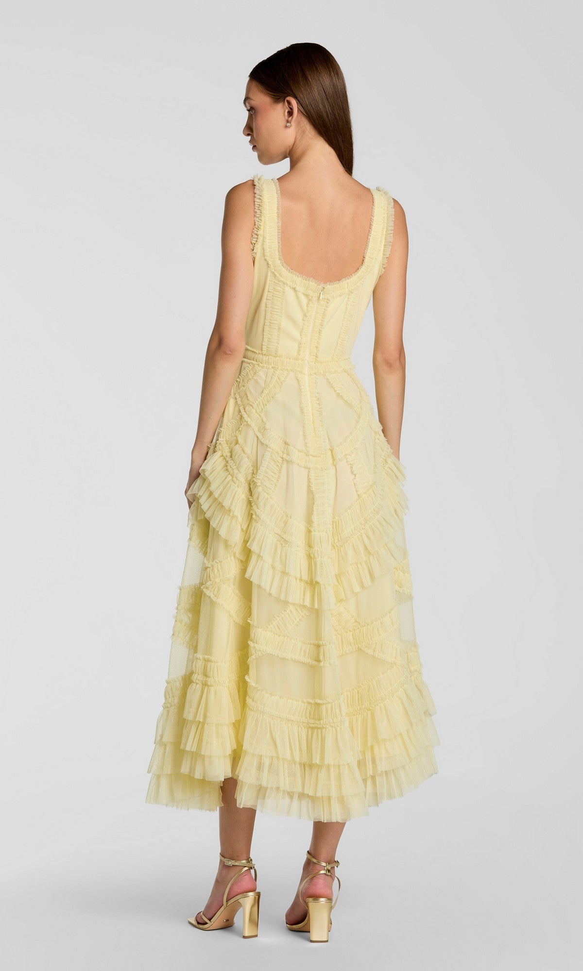 A woman faces away wearing the Mac Duggal 8250 Midi Wedding Guest Dress—a sleeveless, light yellow A-line dress with layered tiers and micro ruffles—paired stylishly with strappy high-heeled sandals.