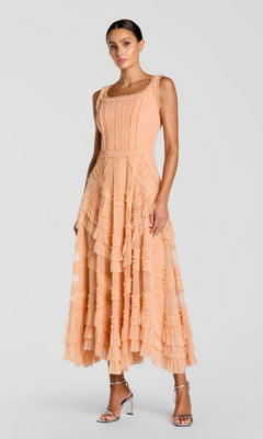A woman in a peach Mac Duggal 8250 midi wedding guest dress with micro ruffle details and silver high-heeled sandals stands against a plain light background.