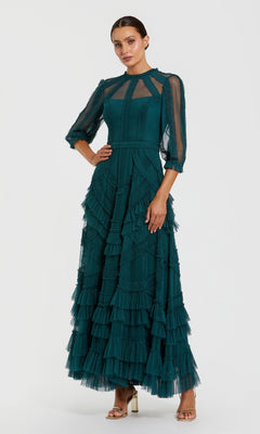 A woman in the Mac Duggal 8251 long formal dress in teal, featuring mesh fabric, sheer sleeves, and layered ruffles, stands in heels against a plain backdrop.