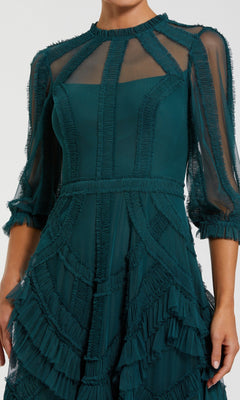 A person wearing the Mac Duggal 8251 long formal dress in dark green, featuring sheer panels, geometric seams, tiered ruffles, and three-quarter sleeves.