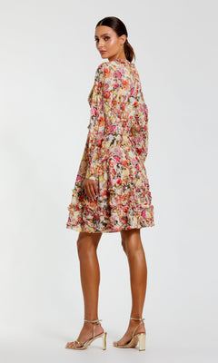 A woman models the Mac Duggal 8261 Micro-Ruffle Wedding-Guest Dress—long-sleeved, knee-length, floral-print with ruffle accents—paired with gold high heels, against a plain white background.