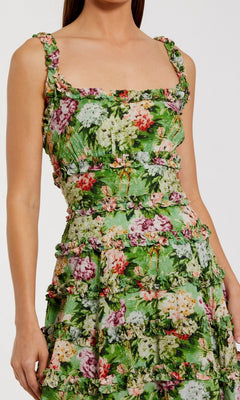 A woman wears the Midi Wedding Guest Dress: Mac Duggal 8263, a sleeveless cotton midi dress with a square neckline, tiered detailing, and a colorful floral print on a green background.
