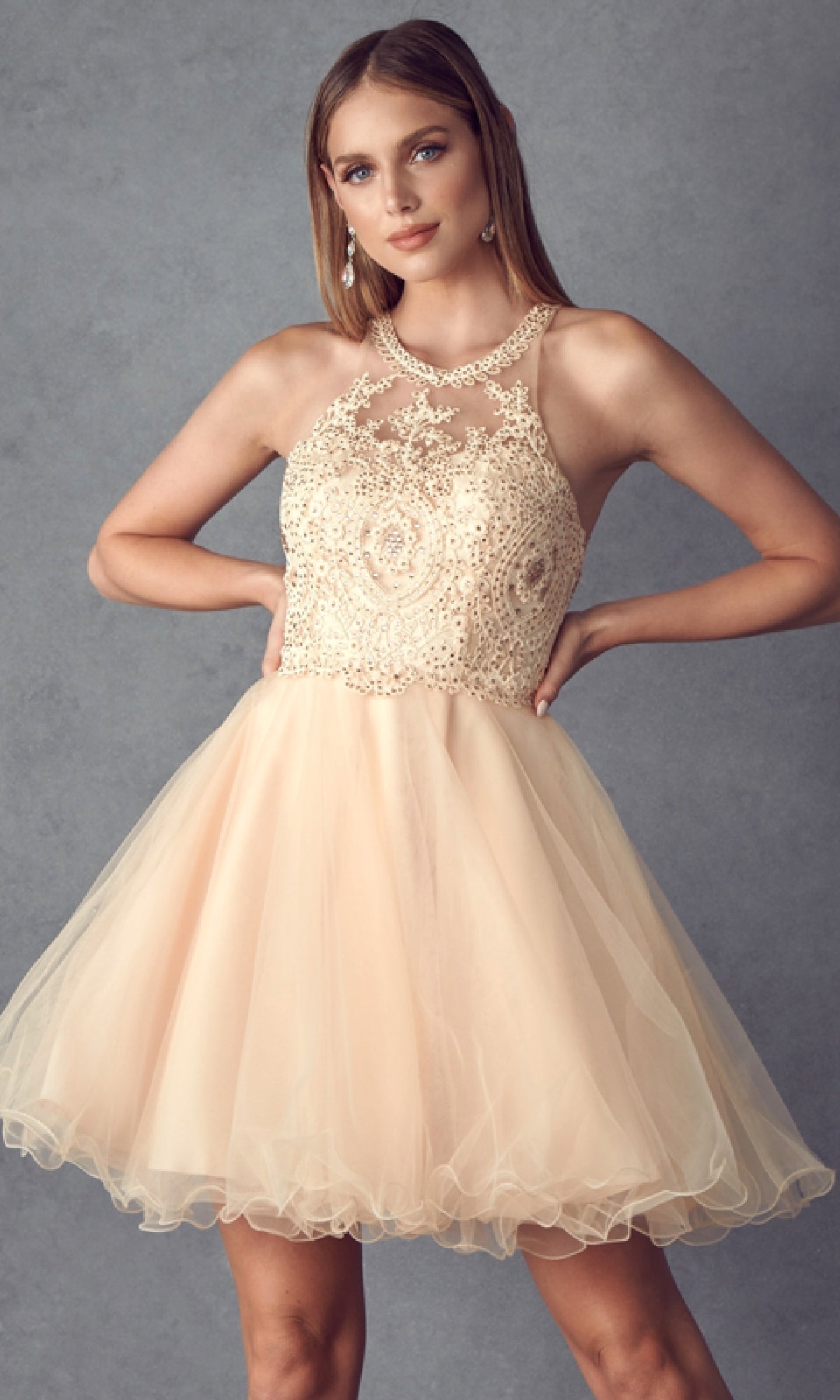 A woman stands against a plain gray background wearing the Short Hoco Dress: Juliet 826—a sleeveless, light beige, knee-length babydoll homecoming dress with a lace bodice and an A-line tulle skirt.