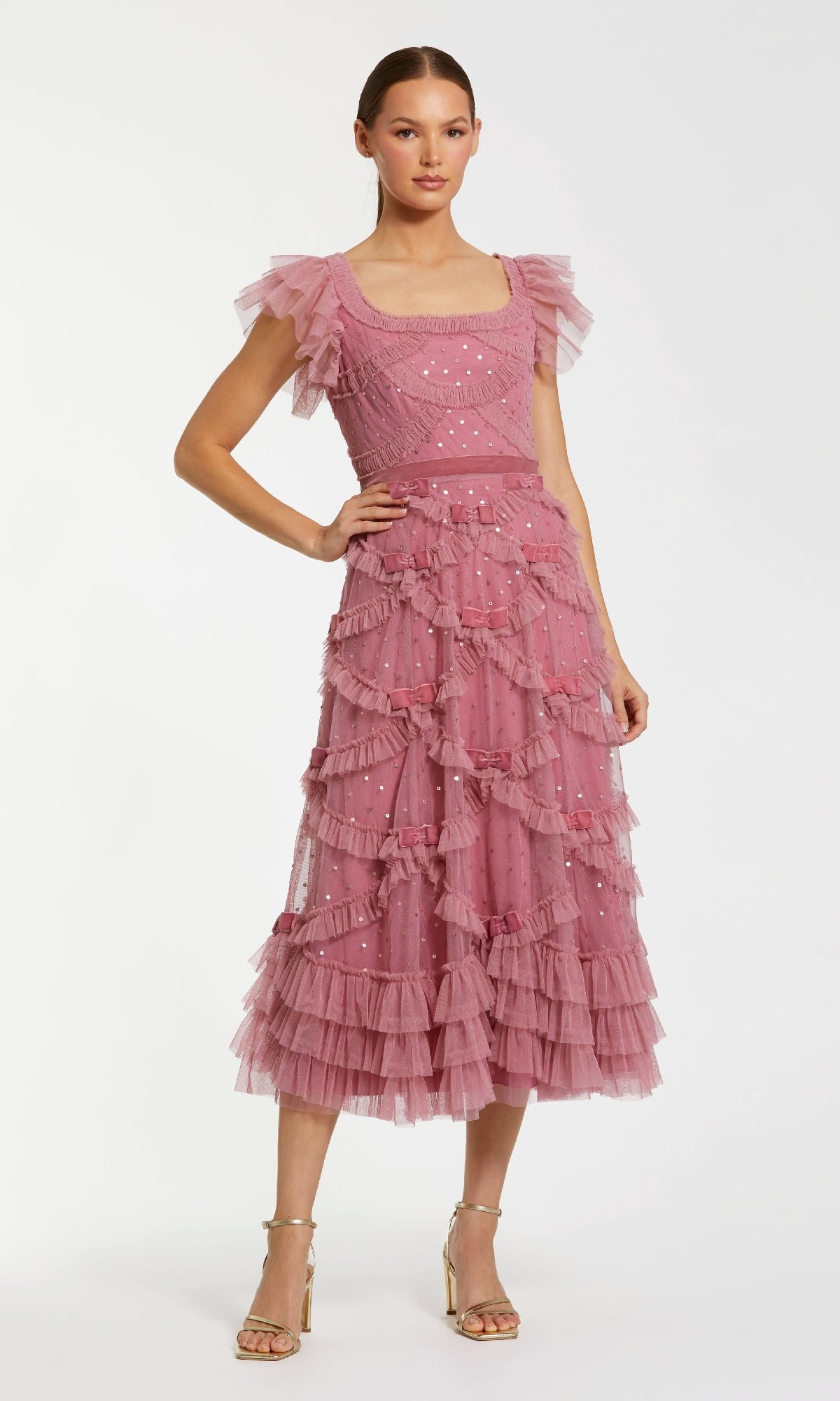 A woman wears the Antique Rose Wedding Guest Dress: Mac Duggal 8272—a ruffled, tea-length tulle dress with sequin accents and flutter sleeves—paired with gold high-heeled sandals against a plain background.