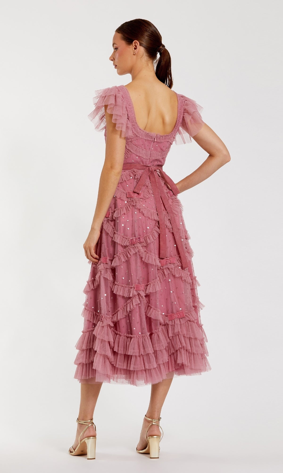 A woman faces away in the Antique Rose Wedding Guest Dress: Mac Duggal 8272, a pink tea-length dress with ruffles, short sleeves, and a bow waist, styled with beige heels—charming for any wedding guest.