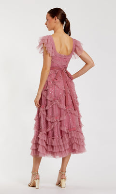A woman faces away in the Antique Rose Wedding Guest Dress: Mac Duggal 8272, a pink tea-length dress with ruffles, short sleeves, and a bow waist, styled with beige heels—charming for any wedding guest.
