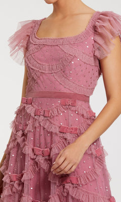 A person wears the Antique Rose Wedding Guest Dress: Mac Duggal 8272, a tea-length pink tulle dress with ruffles, sequin accents, short flutter sleeves, and small bows on the skirt.