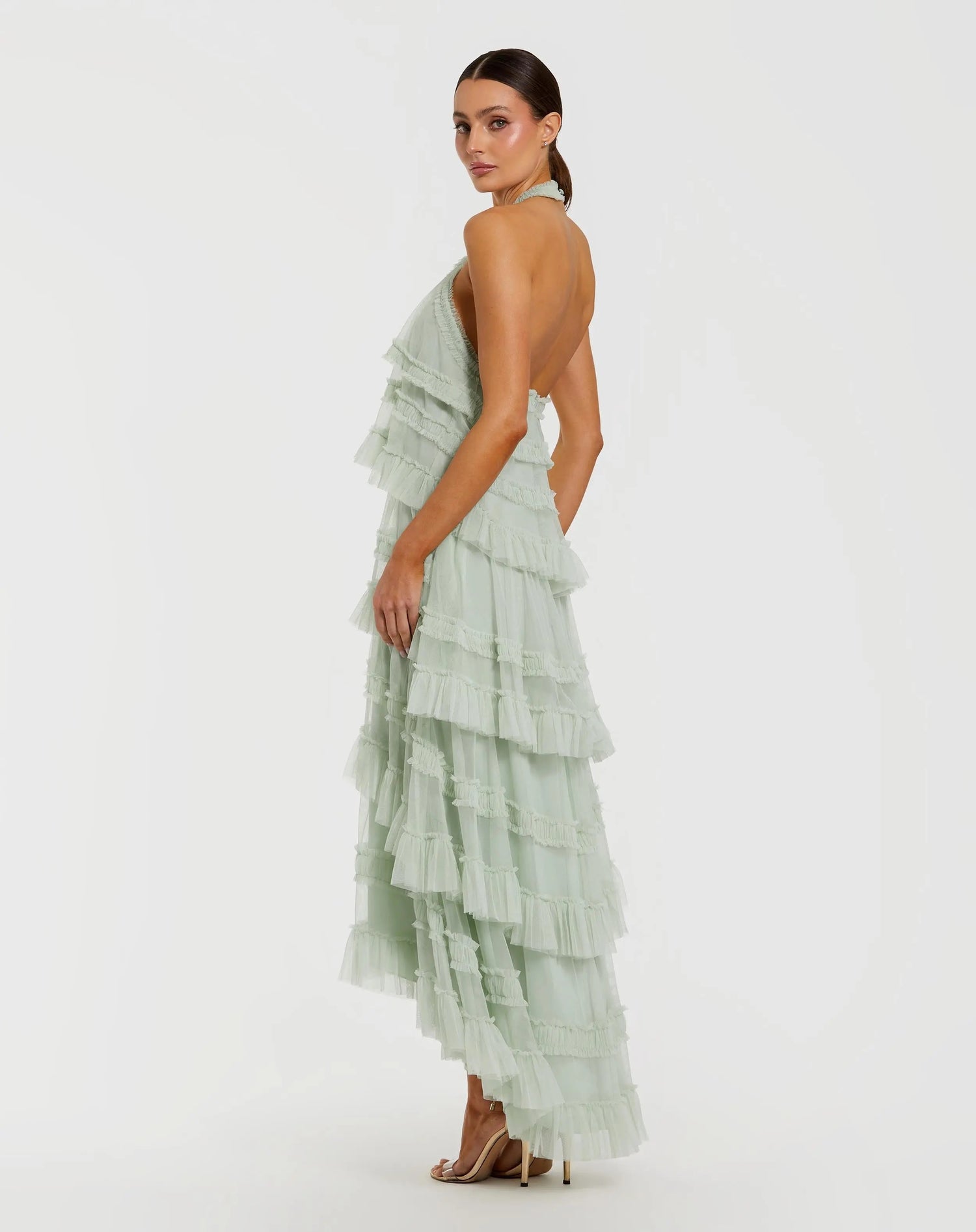 A woman poses sideways against a plain white background, showcasing the elegant tiered layers and high-low hemline of the Mac Duggal 8276 Tea-Length Party Dress in pale green, styled with nude high heels.