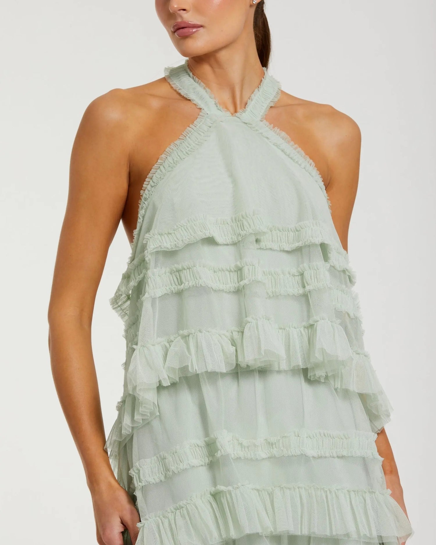 A woman is shown from the shoulders to mid-thigh against a plain background, wearing the Mac Duggal 8276 Tea-Length Party Dress—a light green halter mesh ruffle design with multiple ruffled layers and a stylish high-low hemline.