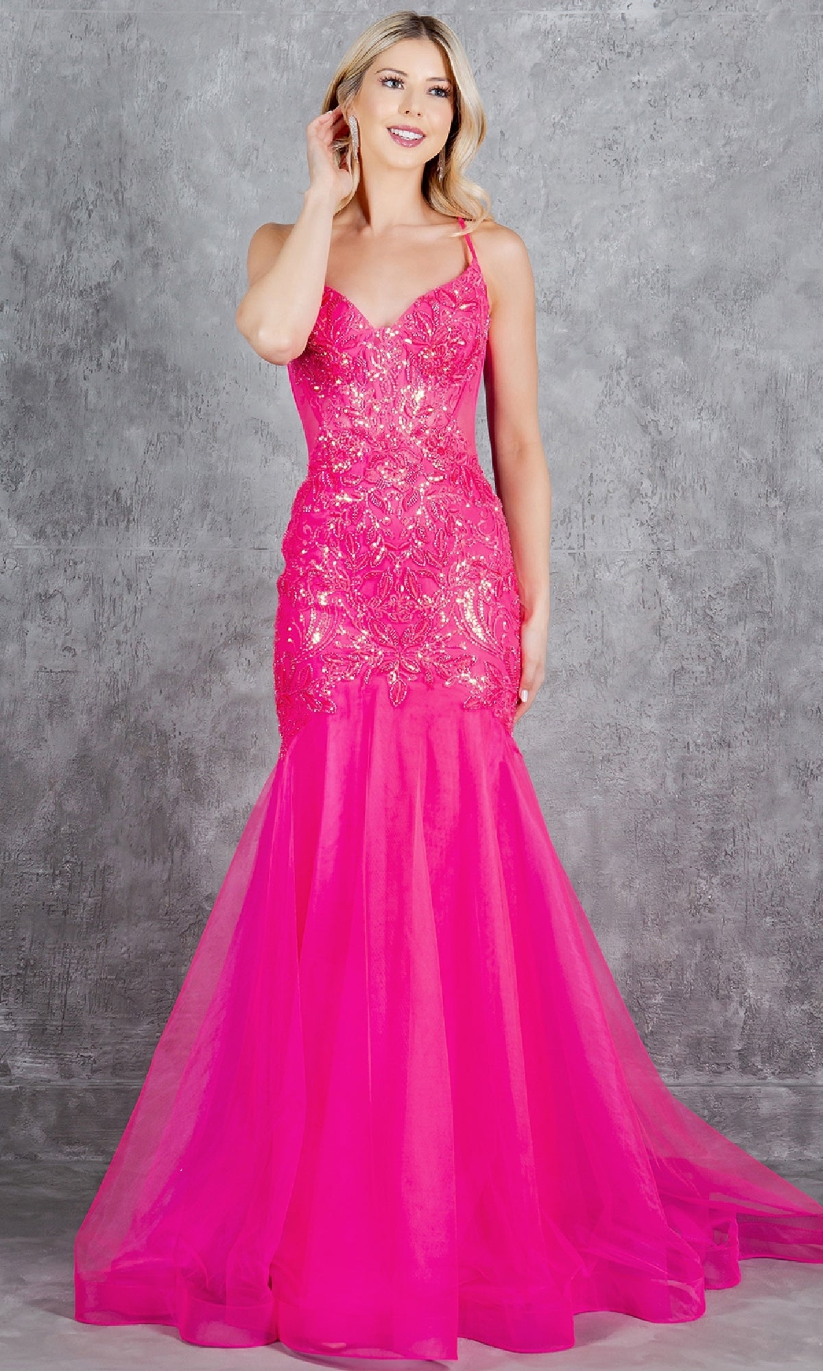 A woman wearing the Cinderella Couture 8276J, a bright pink, sleeveless, sequin mermaid-style prom dress, stands before a gray textured backdrop.