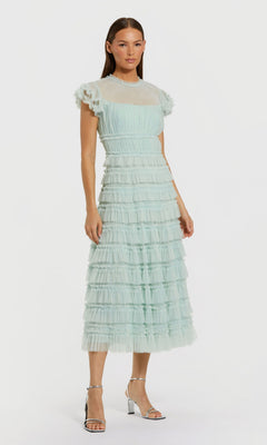 A woman wears the mint Midi Wedding Guest Dress: Mac Duggal 8285 with a sheer high neck, fit-and-flare silhouette, and silver heels, standing against a plain white background.