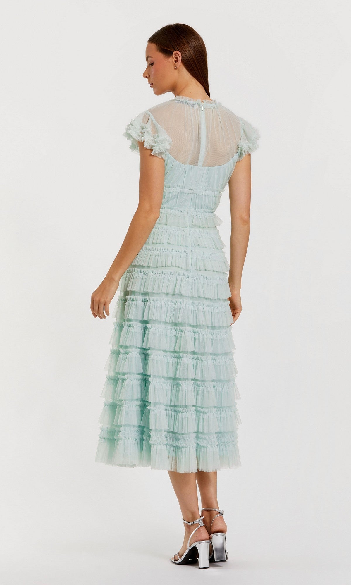 Woman in profile wears the Mac Duggal 8285 Midi Wedding Guest Dress in light blue, featuring a sheer high neck, ruffle details, and a fit-and-flare silhouette, paired with silver heels against a plain background.