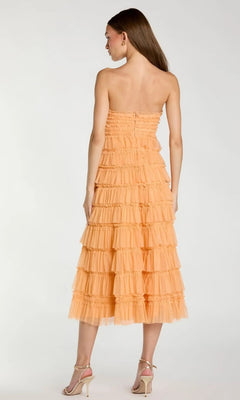 A woman stands facing away, wearing the Midi Semi-Formal Dress: Mac Duggal 8286—a strapless peach wedding guest dress with micro-ruffle detailing—paired with high-heeled sandals.